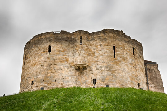 An exterior view of the historic stone Clifford's Tower (York Castle) sitting on a grassy mound under an overcast, moody sky in York, United Kingdom. HDR.