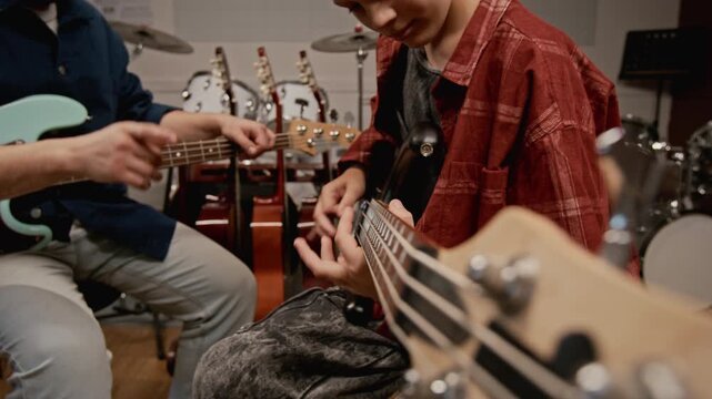Teenager practicing bass guitar during music lesson with young instructor guiding him. Video for music school advertisements or instrument retail promotions