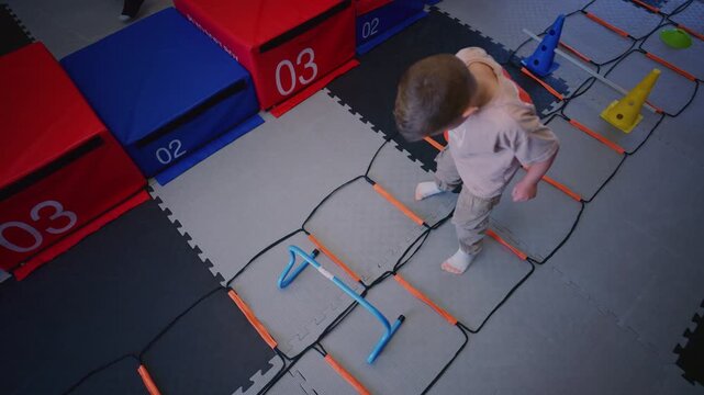 A young child performs agility ladder drills and steps over small hurdles in a dedicated play area. The boy develops motor skills and coordination through engaging kids fitness activities.