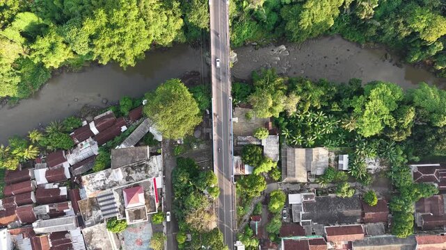 High angle drone photograph of a river crossing and busy road flanked by dense trees and closely packed houses, capturing neighborhood life, tropical vegetation, bridges, and urban