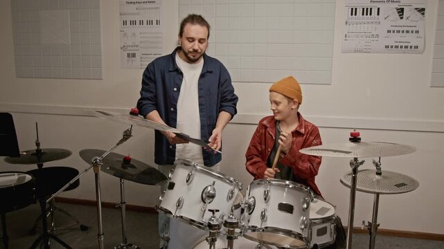 Young male teacher explaining sheet music to teenage boy sitting at drum kit. Footage for music school advertisements and youth education campaigns