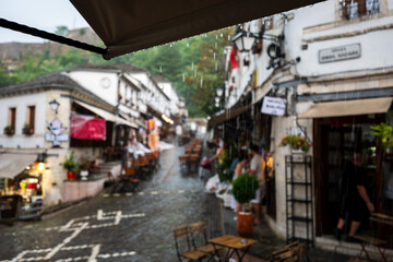 Fototapeta premium Rain falling over charming old town street with cafés and traditional architecture, creating a cozy atmosphere in a historic European destination in Gjirokastra, Albania