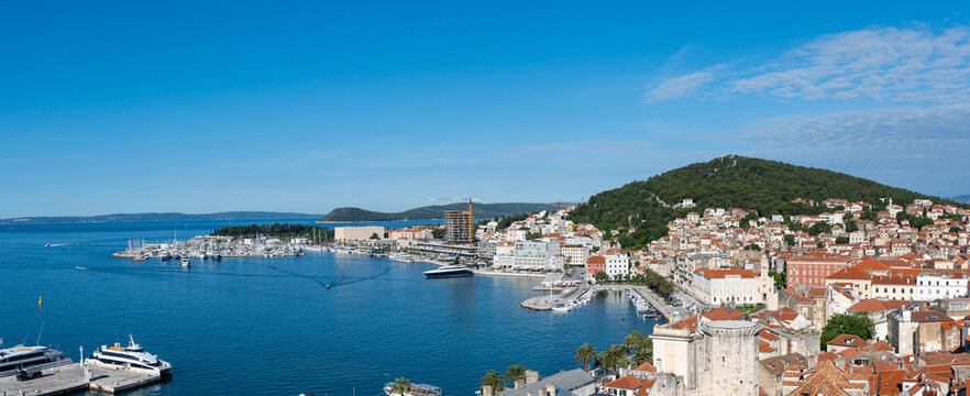 Wide panoramic view of Split, Croatia, featuring the historic old town, waterfront marina, luxury yachts, Adriatic Sea, and Marjan Hill beneath a bright blue sky.