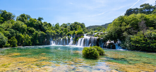 Crystal-clear turquoise water flows over cascading waterfalls in Krka National Park, Croatia, surrounded by lush green forest under a bright summer sky. © OlgaPS