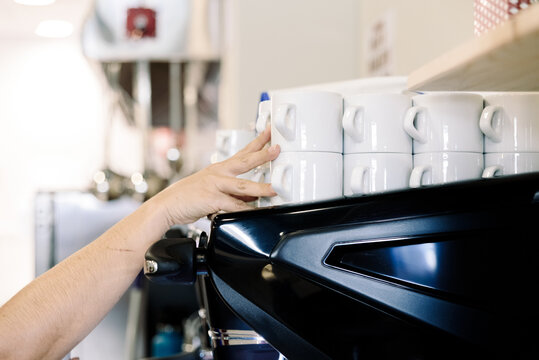 A worker is placing clean coffee cups in order on a cafe counter. The scene shows a busy bar environment in the morning