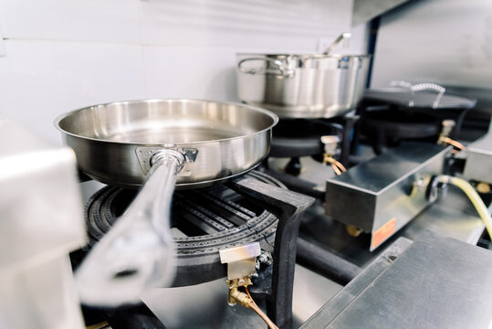 Steel pots and pans sit on gas burners in a professional kitchen, prepared for cooking various meals during a busy service