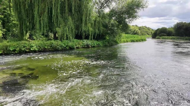 Fullerton Hampshire England UK. 29.06.2026. Video. A tranquil river winds its way through verdant foliage, with weeping willows lining the banks under a dramatic sky