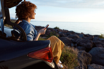 Fototapeta na wymiar Young woman with curly hair sits in a car trunk by a rocky shore, scrolling on a smartphone as the sun sets over the calm sea during golden hour