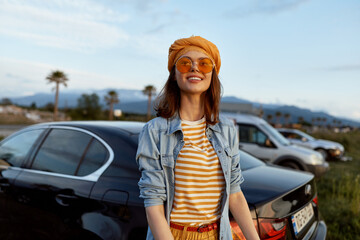 Naklejka premium Smiling woman outdoors wearing striped shirt denim jacket orange hat and sunglasses. Casual lifestyle portrait in bright daylight with a relaxed, cheerful mood near parked cars.