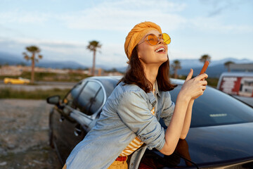 Naklejka premium woman lifestyle outdoors smiling, sunglasses car sunny casual scene of a cheerful woman leaning on a car in bright sun, denim jacket and headscarf fluttering in breeze