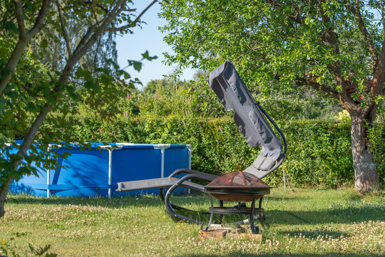 A sunny backyard contains an above ground pool a reclining chair and a metal fire pit. Mature garden trees shrubs and a green hedge surround the lawn.