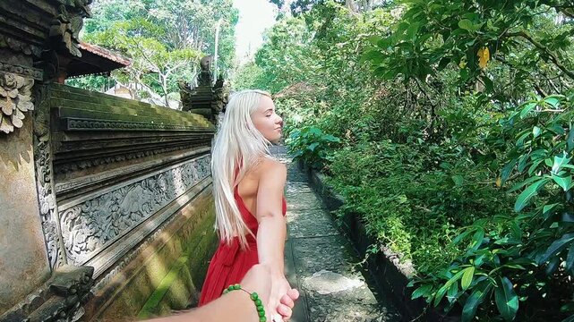 Blonde woman in red backless dress moves along narrow stone path beside carved temple wall, holding joined hands, turning back with smile, framed by lush tropical foliage and sunlit garden calm