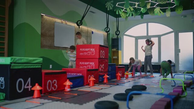 A group of diverse children participates in an active fitness class, navigating various obstacles in a colorful gym.
