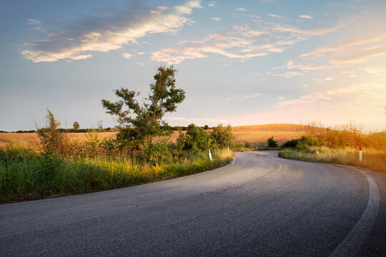 Winding country road through golden field at sunset, curvy asphalt road landscape in Tuscany, Italy