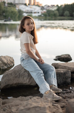 A young girl with long brown hair sits on a rock by the water, wearing a casual outfit and enjoying the serene lake view.