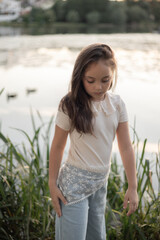 A thoughtful young girl with long brown hair stands by a lake, wearing a light sweater and flowy blue pants, surrounded by greenery. © VAshowcase