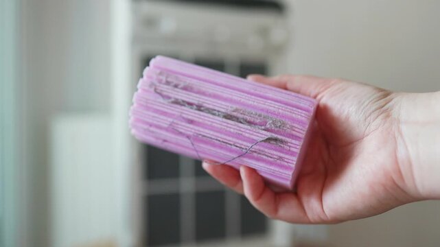 A close-up shot of a young woman hands removing or installing a square black mesh dust filter from a white modern household air purifier in a bright room.