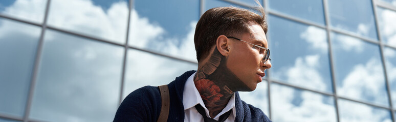 A young man with tattoos and stylish glasses stands against a glass building reflecting clouds. © LIGHTFIELD STUDIOS