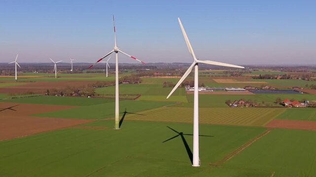Aerial wide angle view of multiple tall wind turbines generating clean energy over green farmlands and rural agricultural countryside fields under a clear blue sky on a bright sunny day