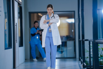Fototapeta premium Nursing care: Confident young Asian female doctor in a white coat and stethoscope walking through a hospital corridor at night during her shift, warm caring atmosphere