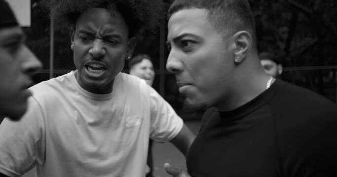 Heated argument between two men of Latin and African descent on outdoor basketball court, face to face in conflict as teammate steps in to defuse tension, black and white image