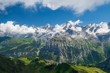 Fototapeta premium Wide view of Swiss Alps above Lauterbrunnen valley, with green slopes, rocky ridges, lingering snowfields, and summer clouds under blue sky.