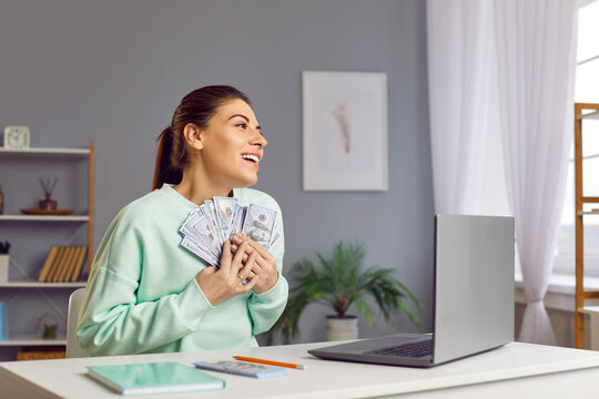 Happy woman sits at a table at home, with laptop, holding stack of cash, money in hands. She celebrating success, receiving a salary or winning a prize. Financial achievement and wealth.