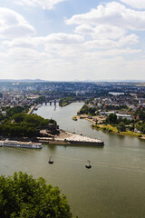 Obraz premium Elevated panoramic view of Koblenz with the Rhine and Moselle river confluence, Deutsches Eck, cable cars, bridges, and historic architecture beneath a dramatic summer sky.