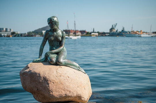 Copenhagen Denmark - August 13 2025. The Little Mermaid statue is silhouetted against Copenhagen harbour with boats and the waterfront behind her the famous bronze figure on her rock at the