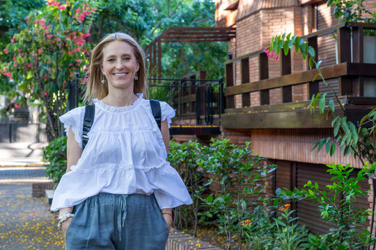 Portrait ofa a beautiful lady wearing a backpack and wide-leg trousers posing while walking towards the camera in a green suburban setting