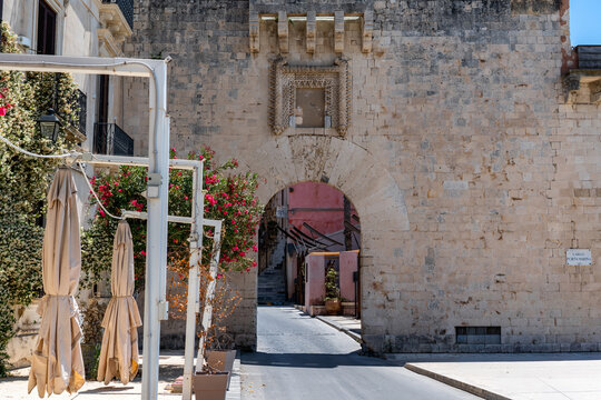 Porta Marina in Syracuse, a 15th-century Spanish stone arch leading into the medieval district of Ortigia, surmounted by an elegantly carved Catalan-style shrine featuring floral motifs 