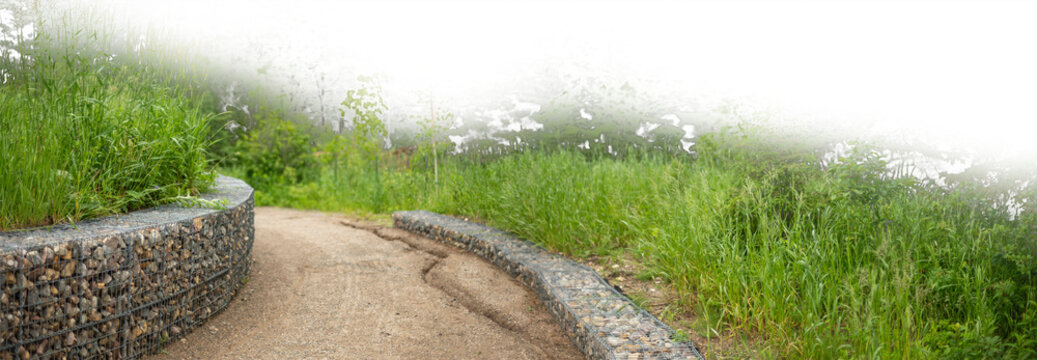 A curving gravel walking path with gabion walls and a partially transparent sky
