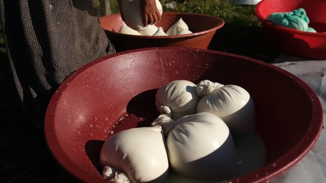 A woman squeezes a cloth bag of fresh curds over a red basin, showing a traditional cheese-making stage, manual whey draining, rural food preparation, and small-scale outdoor dairy production by hand.