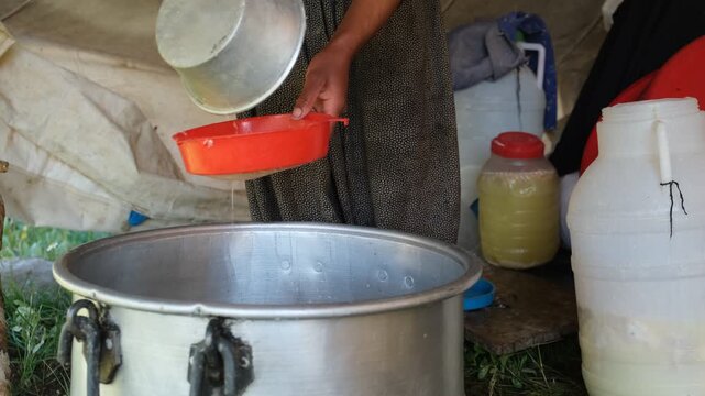 A pale dairy mixture is poured through a strainer into a metal pot during traditional cheese production, capturing manual filtration, rural food preparation, and small-scale dairy processing outdoors.