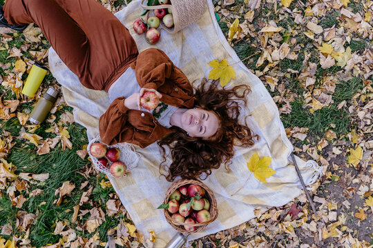 Thoughtful red haired woman holding fresh apple during autumn picnic in orchard