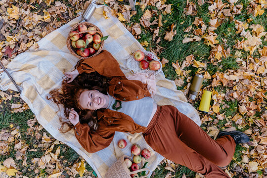 Relaxed red haired woman resting among fresh apples during cozy autumn orchard picnic