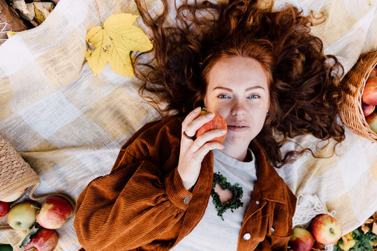 Serene freckled red haired woman holding ripe apple during cozy autumn orchard picnic