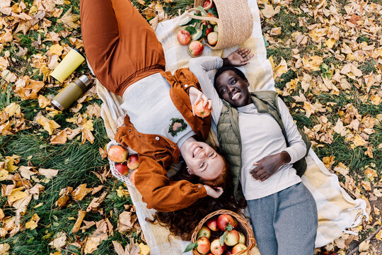 Joyful diverse female friends relaxing together after apple harvest during autumn orchard picnic