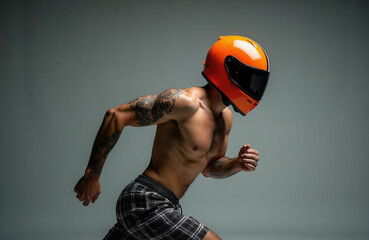 Obraz premium Muscular male athlete with arm tattoo wearing orange motorcycle racing helmet runs in studio. Shirtless fit guy dressed in plaid shorts moves fast like pro kart driver. Dynamic motion shot of