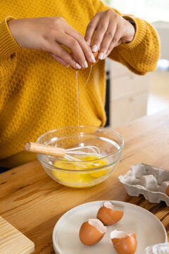 African American woman cracking egg into mixing bowl in yellow sweater at wooden table with whisk