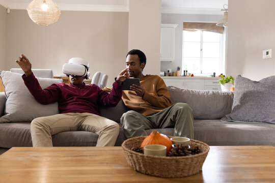 African American father and teenage son sitting on grey sofa wearing VR headset holding tablet