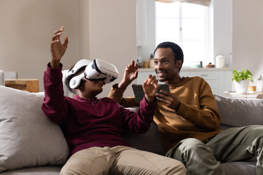 African father holding phone and teenage son sitting on gray sofa wearing white VR headset reacting