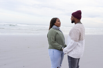 Obraz premium Couple holding hands, facing each other on wet sand at shoreline, wearing knit sweaters, watch