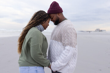 Obraz premium African American couple standing touching heads and holding hands on beach wearing beanie, sweaters