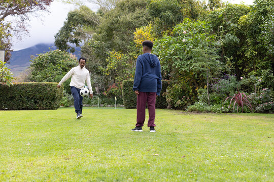 African American father and teenage son playing soccer with ball on garden lawn wearing hoodies