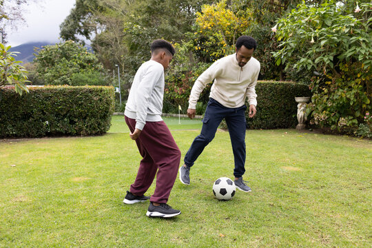 Man and teen male kicking soccer ball on backyard lawn near trimmed hedges and urn