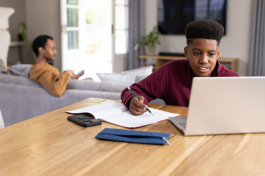 Dad and teenage son at home, son studying at table with laptop while dad scrolling smartphone