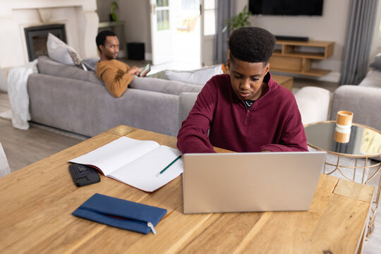 African American father and teenage son working at wooden dining table in living room with laptop