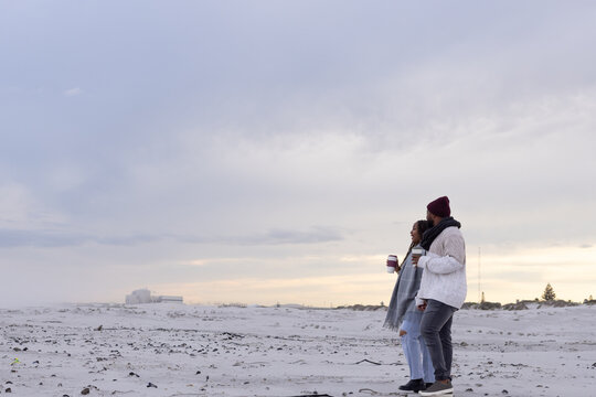 African American couple walking along sandy shoreline in coats holding takeaway cups, copy space