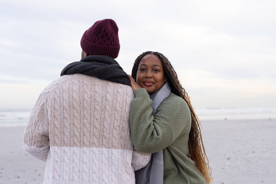 Couple standing close on sandy beach, woman resting hand on man's shoulder, wearing knit scarves